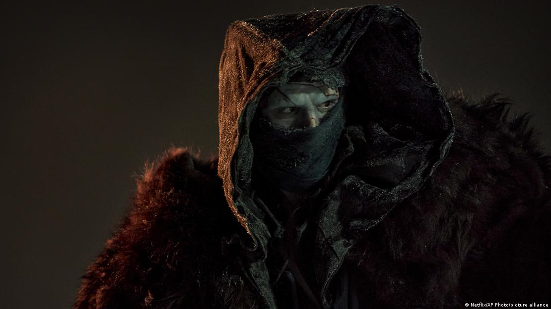 Film still from 'Frankenstein': The Creature, a character wearing a rugged fur coat, face covered up to the eyes with a dark scarf.