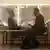 Two men wearing suits sit in front of a mirror and laugh at each other