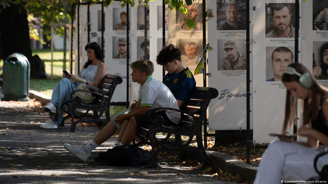 Junge Menschen sitzen in Lwiw auf Parkbänken vor dem Hintergrund aufgehängter Portraits gefallener Soldaten