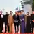 8 people wearing dresses and suits stand on a red carpet