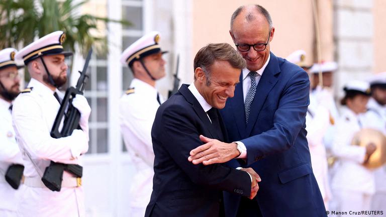 Macron ve Merz tokalaşıyorlar. Merz boşta kalan eliyle Macron'un omzuna dostça dokunuyor