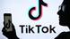 Persona sostiene celular con el símbolo de TikTok en pantalla. Al fondo, un gran cartel con el logo y las palabras TikTok. Persona sostiene celular con el símbolo de TikTok en pantalla. Al fondo, un gran cartel con el logo y las palabras TikTok.