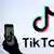 Persona sostiene celular con el símbolo de TikTok en pantalla. Al fondo, un gran cartel con el logo y las palabras TikTok.