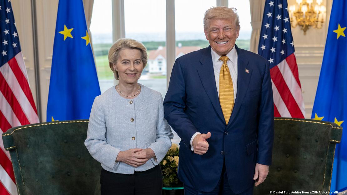 Von der Leyen und Trump; von der Leyen hat die Hände gefaltet, Trump grinst und hält einen Daumen hoch, im Hintergrund EU- und USA-Flaggen