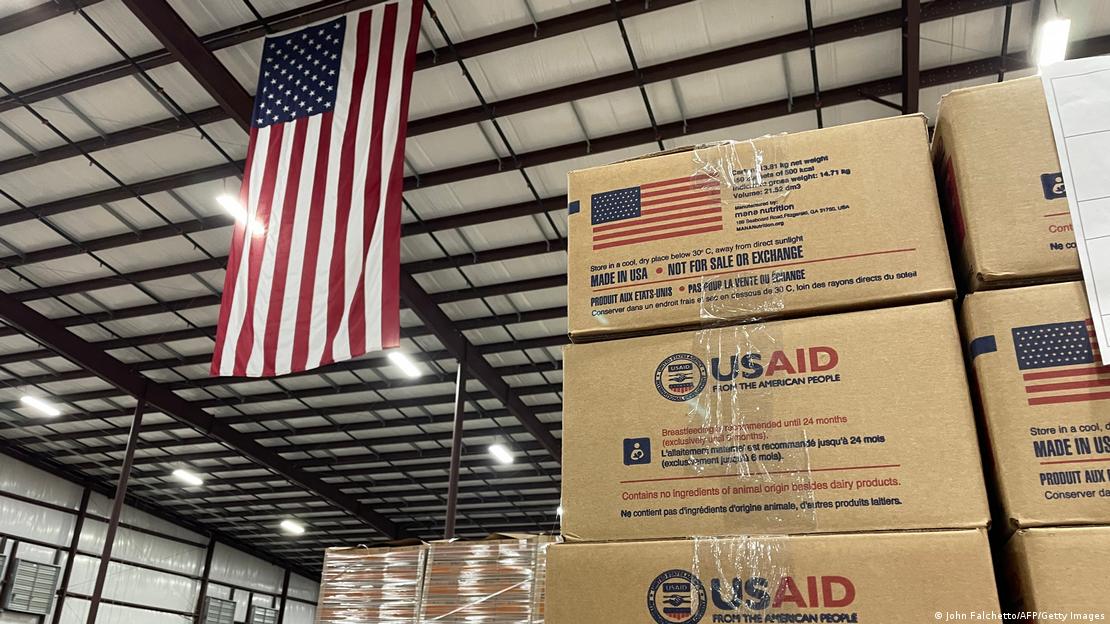 Kisten mit der Aufschrift USAID, dahinter eine große Flagge der USA