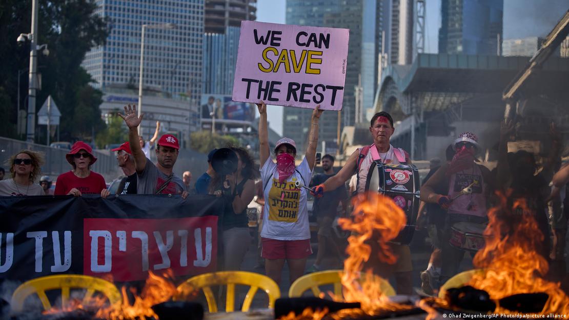 Manifestantes atrás de barricada em chamas protestam bloqueando avenida e pedindo libertação dos reféns; mulher segura cartaz com dizeres "podemos salvar o restante"