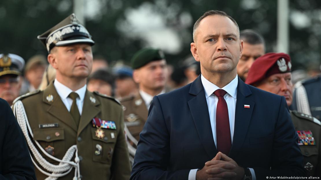Ein Mann im dunklen Anzug, Polens Präsident Nawrocki, steht mit gefalteten Händen neben einem Offizier in Uniform. Hinter den beiden sind weitere Uniformierte zu sehen