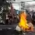 Protesters, some with red flags, stand behind a pile of burning cardboard