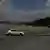 A car drives down a road, a tall grey wall is seen in the background