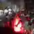 Varios manifestantes golpean los escudos protectores de los policías antimotines mientras se ve algo arder en el suelo.