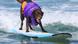 Ein brauner Labrador mit lila Trikot und Schwimmweste steht auf einem Surfboard, das auf den Wellen treibt. Ein brauner Labrador mit lila Trikot und Schwimmweste steht auf einem Surfboard, das auf den Wellen treibt.