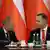 Two men (Donald Tusk and Karol Nawrocki) sit at a table in front of a row of Polish flags. Tusk looks intently at Nawrocki, who appears to be speaking
