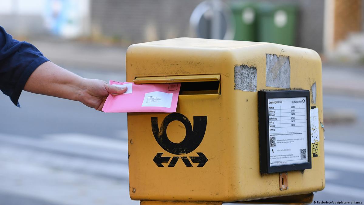 A person drops a pink postal voting envelope into a yellow mailbox in Germany