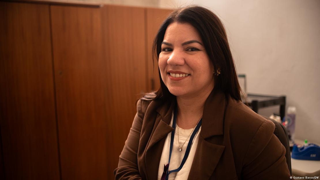 A agente social Amanda Lourenço, de cabelos castanhos e lisos e vestida com blazer marrom, sorri para a câmera diante de armário de madeira em escritório.
