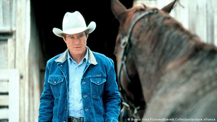 Robert Redford in a denim jacket and white cowboy hat,standing in front of a horse.