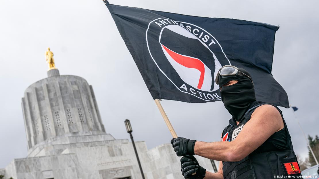 Ein Demonstrant mit schwarzen Handschuhen und schwarzer Sturmhaube trägt eine antifaschistische Flagge 