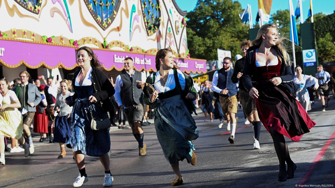 Tres mujeres jóvenes, ataviadas con el traje tradicional bávaro, corren en primer plano seguidas de una multitud, tras acceder al recinto ferial.