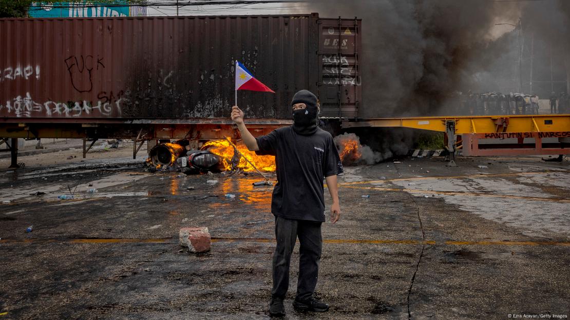 Filipino com rosto coberto por balaclava segura bandeira filipina em frente à barricada com um contêiner em chamas