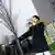 person with dark hair and a long jacket plays a short trumpet while standing on the street. 