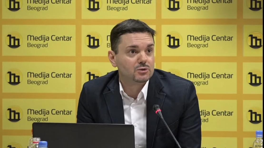 Mlađi muškarac na pozornici pred mikrofonm , iza njega žuti zid s natpisima