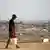 In the foreground an out of focus man carrying a bucket of water, in the background rubble of buildings in Gaza