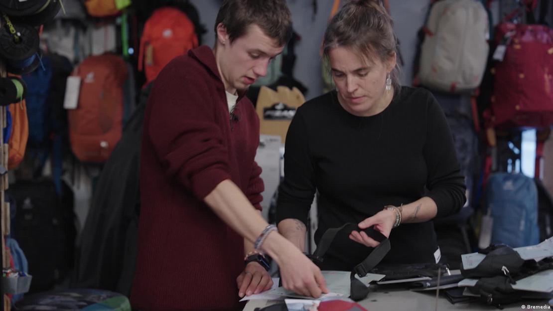 Un joven (17 años) habla con una compañera detrás del mostrador de la tienda sobre material para actividades al aire libre.