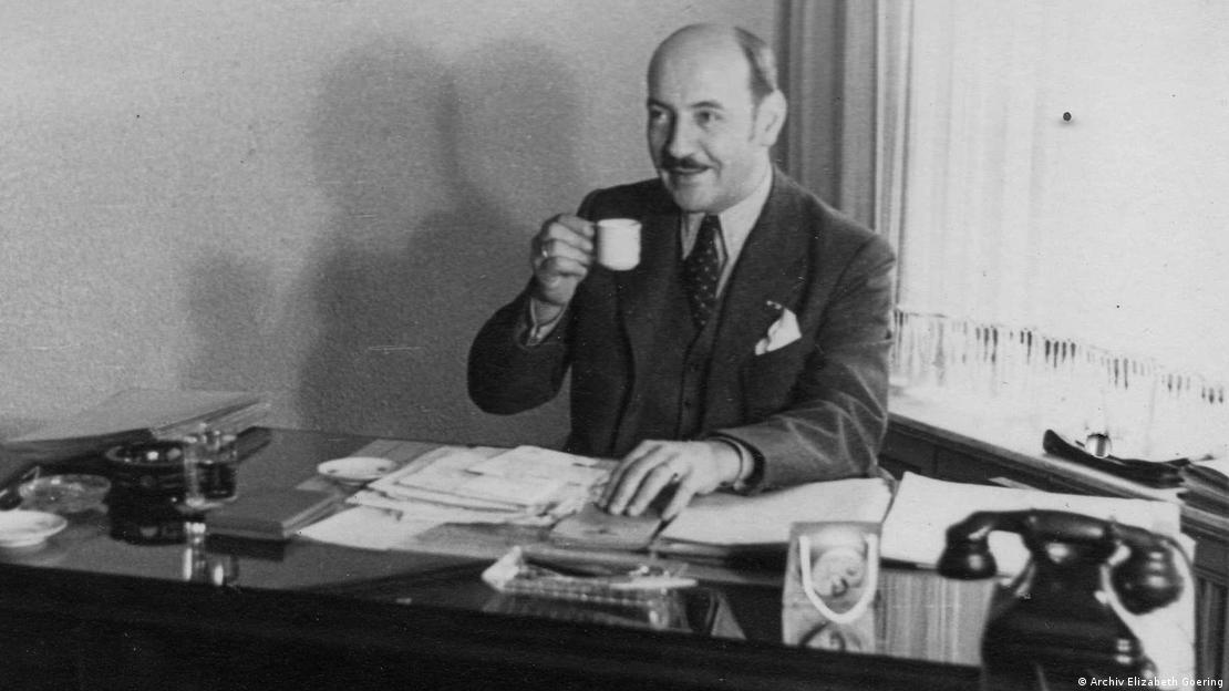 Black and white picture of a smiling man in suit, holding up a cup while seated at a desk.