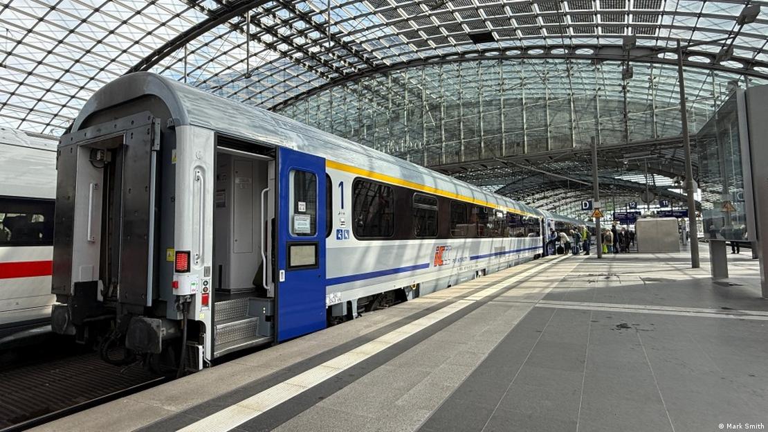 A train with yellow and blue design is standing at a large train station under a big roof, some people are about to enter a compartement.