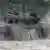 Ein deutscher Leopard 2 Panzer fährt durch Matsch, Dreck spritzt hoch, im Hintergrund Büsche.