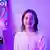A young woman with shoulder-length brown hair, wearing a simple white button-up shirt with a collar, looking at the camera. She is lit by a purple light and sits next to a feathery lamp and a colorful wall hanging. 