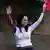 María Corina Machado, con las manos hacia arriba, ondea con su mano izquierda una bandera de Venezuela.