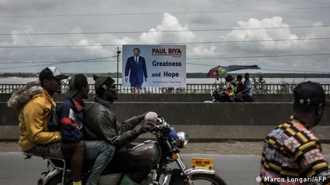 Des navetteurs empruntent des motos-taxis pour traverser un pont devant une affiche électorale représentant le président camerounais sortant Paul Biya à Douala, le 7 octobre 2025