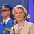 Eine Frau in beigefarbenem Hosenanzug (Ursula von der Leyen) blickt leicht an der Kamera vorbei. Im Hintergrund sind unscharf eine EU-Fahne sowie ein Mann in einer blauen Uniform zu sehen