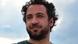 Close-up portrait of a bearded man with curly hair looking to the side, set against a cloudy sky background Close-up portrait of a bearded man with curly hair looking to the side, set against a cloudy sky background