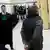 Ein Mann mit einer Base Cap in einem Raum von hinten (Wolodymyr Z.), hinter ihm ein Polizist, schräg vor ihm ein Mann in Robe, im Hintergrund Journalisten mit Kameras