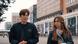 A young man in dark jacket walking past a line of modern buildings, beside a young woman with long brown hair, who is looking at him A young man in dark jacket walking past a line of modern buildings, beside a young woman with long brown hair, who is looking at him