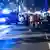 La imagen muestra a varias personas en el lugar de la balacera, entre ellos policías y socorristas, y una ambulancia al fondo.