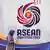 A woman walks past a wall with the ASEAN 2025 logo. She is wearing a headscarf and looking at her phone.