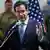 A man in a blue suit (US Secretary of State Marco Rubio) gestures while standing in front of an American flag as soldiers stand behind him