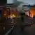 Un bombero observa desde el suelo cómo su compañero lanza un chorro de agua desde el coche escala con el almacén destruido delante.