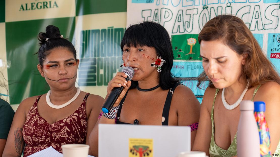 Eine Pressekonferenz von drei indigenen Frauen auf einem Podium. Dahinter ein Plakat. eine Frau mit schwarzen Haaren und bunten Ohrringen spricht. Sie hat Bemahlungen im Gesicht und einen dramatischen Gesichtsausdruck.
