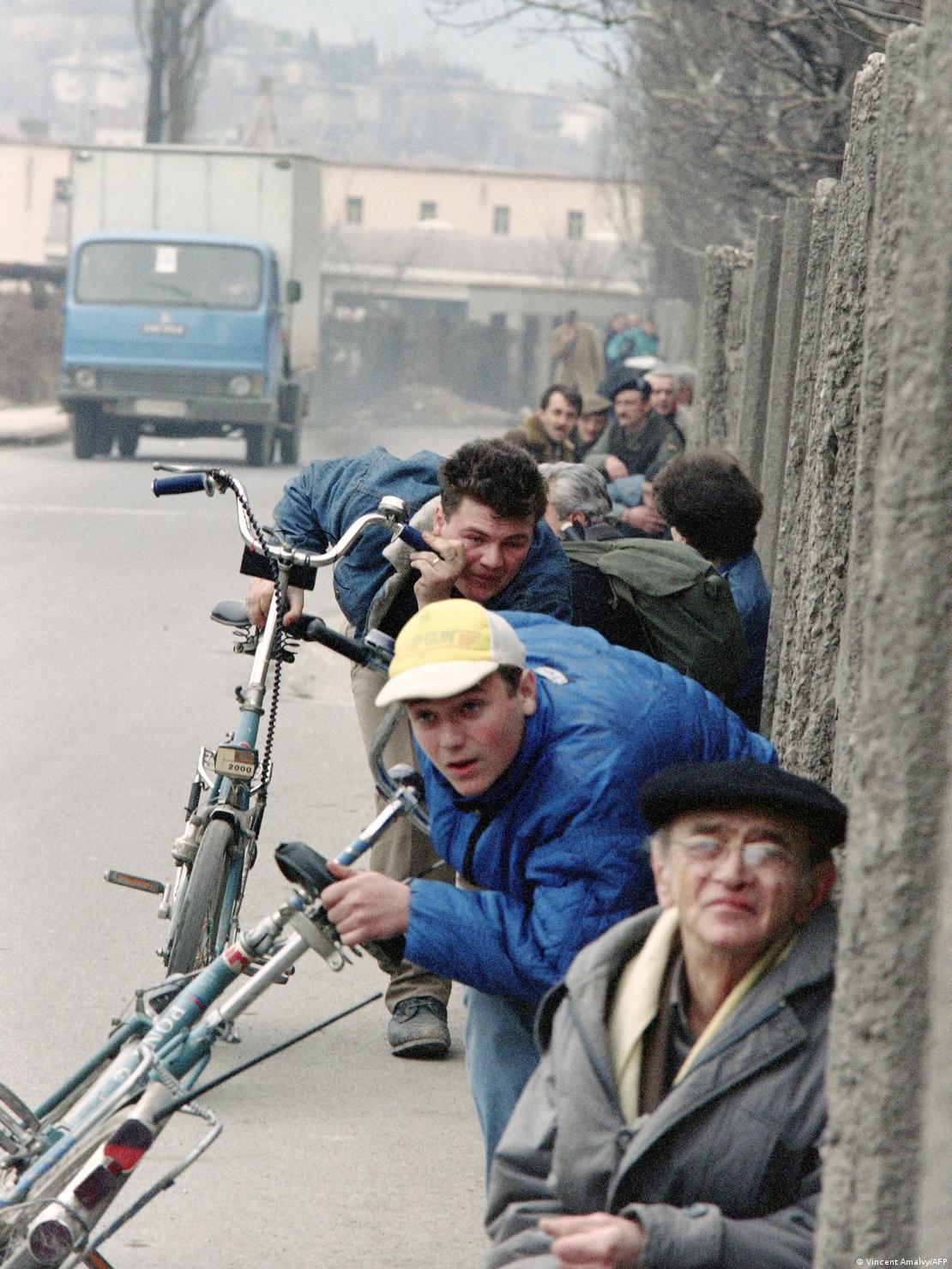 Ljudi prislonjeni uz zid, stari i mladi sa biciklima, traže zaštitu od snajpera