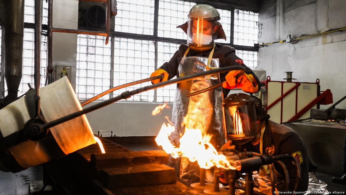 Metal eritme atölyesinde çalışan, koruyucu elbiseler içinde iki işçi görülüyor