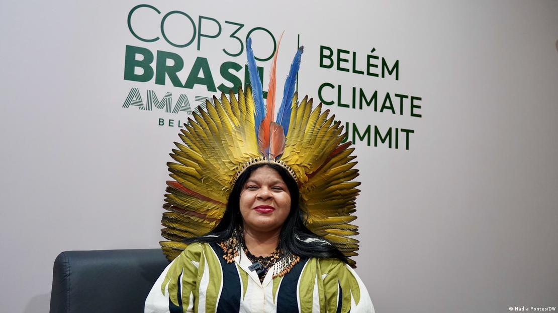 Retrato de la ministra brasileña, ataviada con indumentaria tradicional indígena, que mira sonriente a la cámara con el logo de la COP30 detrás.
