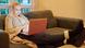 Eine junge blonde Frau sitzt in eine Decke einkuschelt auf dem Sofa und schaut in ein Laptop auf ihren Beinen, die auf dem Couchtisch liegen Eine junge blonde Frau sitzt in eine Decke einkuschelt auf dem Sofa und schaut in ein Laptop auf ihren Beinen, die auf dem Couchtisch liegen