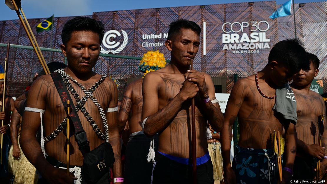Varios miembros del movimiento indígena Munduruku Ipereg Ayu, ataviados con sus trajes y pinturas tradicionales, además de con arcos y flechas, protestan frente al edificio donde se celebra la Cumbre del Clima de la ONU en Belém, con los logos de Naciones Unidas y de la COP30 visibles en la fachada de la sede, así como la bandera de Brasil.