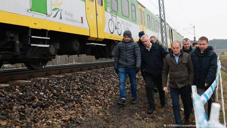 Der polnische Ministerpräsident Donald Tusk und Innenminister Marcin Kierwinski und weitere Männer gehen neben einem stehenden Zug ein Gleisbett entlang, das mit blau-weiße, Flatterband abgesperrt ist