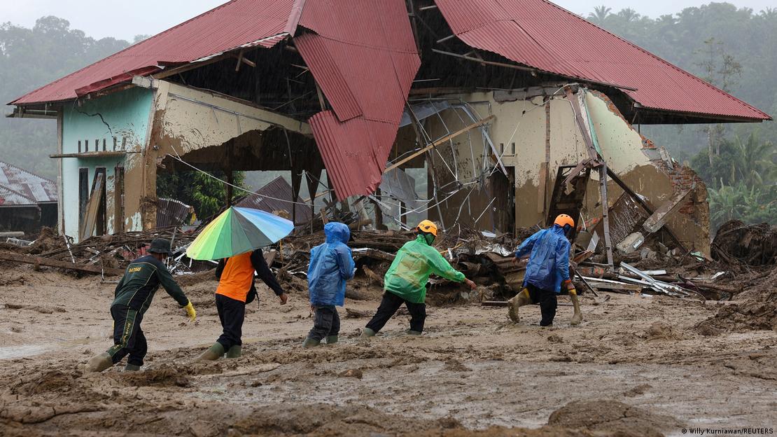 Indonesien Palembayan 2025 | Fünf Männer waten durch tiefen Schlamm, dahinter ein zerstörtes Gebäude 
