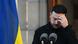 Zelenskyy standing in front of microphones, holding one hand to his head. A Ukrainian flag is next to him. Zelenskyy standing in front of microphones, holding one hand to his head. A Ukrainian flag is next to him.