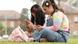 Two young girls sit on the grass as they look at their phones, shown in a side on profile Two young girls sit on the grass as they look at their phones, shown in a side on profile